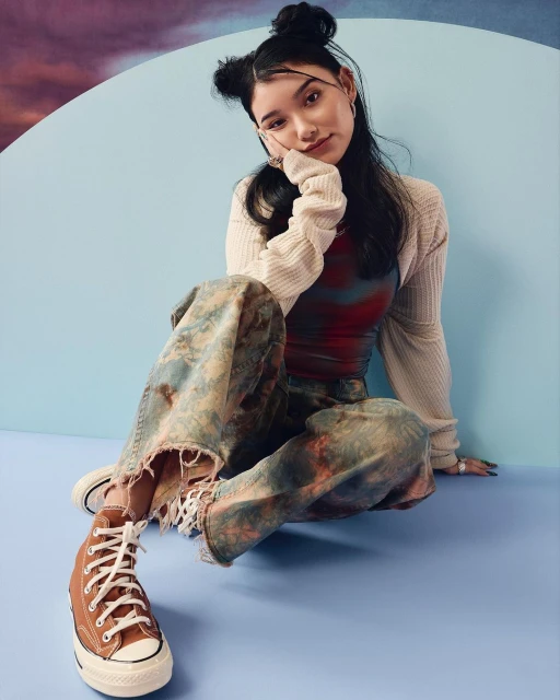 Young adult woman in stylish casual attire posing indoors, wearing tie-dye pants and high-top sneakers, with soft studio lighting.