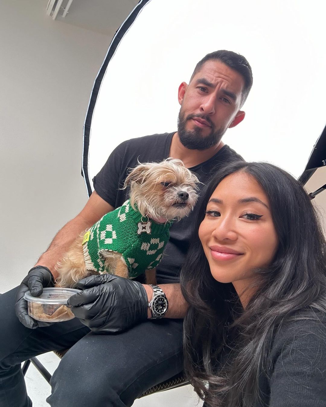 Three people and a dog in a green sweater pose in a bright photo studio, with one person holding a container of dog food.