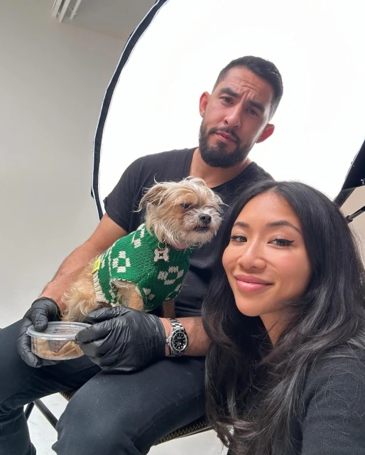 Three people and a dog in a green sweater pose in a bright photo studio, with one person holding a container of dog food.