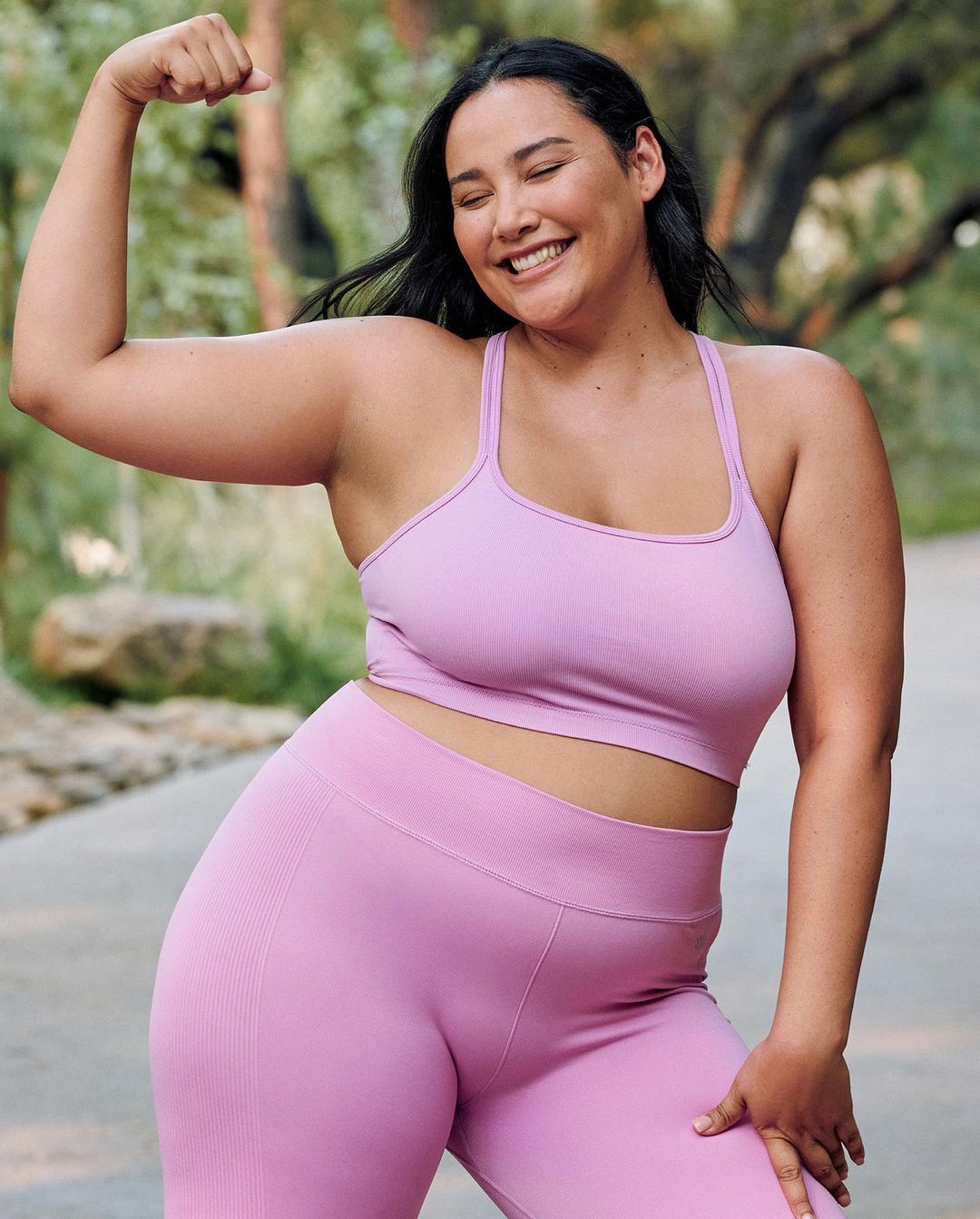 Smiling adult woman in exercise clothing flexes arm muscle outdoors against a green tree backdrop in a portrait-style fitness shot.
