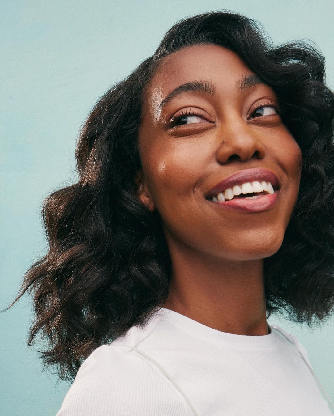 Smiling African-American woman with shoulder-length wavy black hair, wearing white top against a light-blue background in studio lighting.