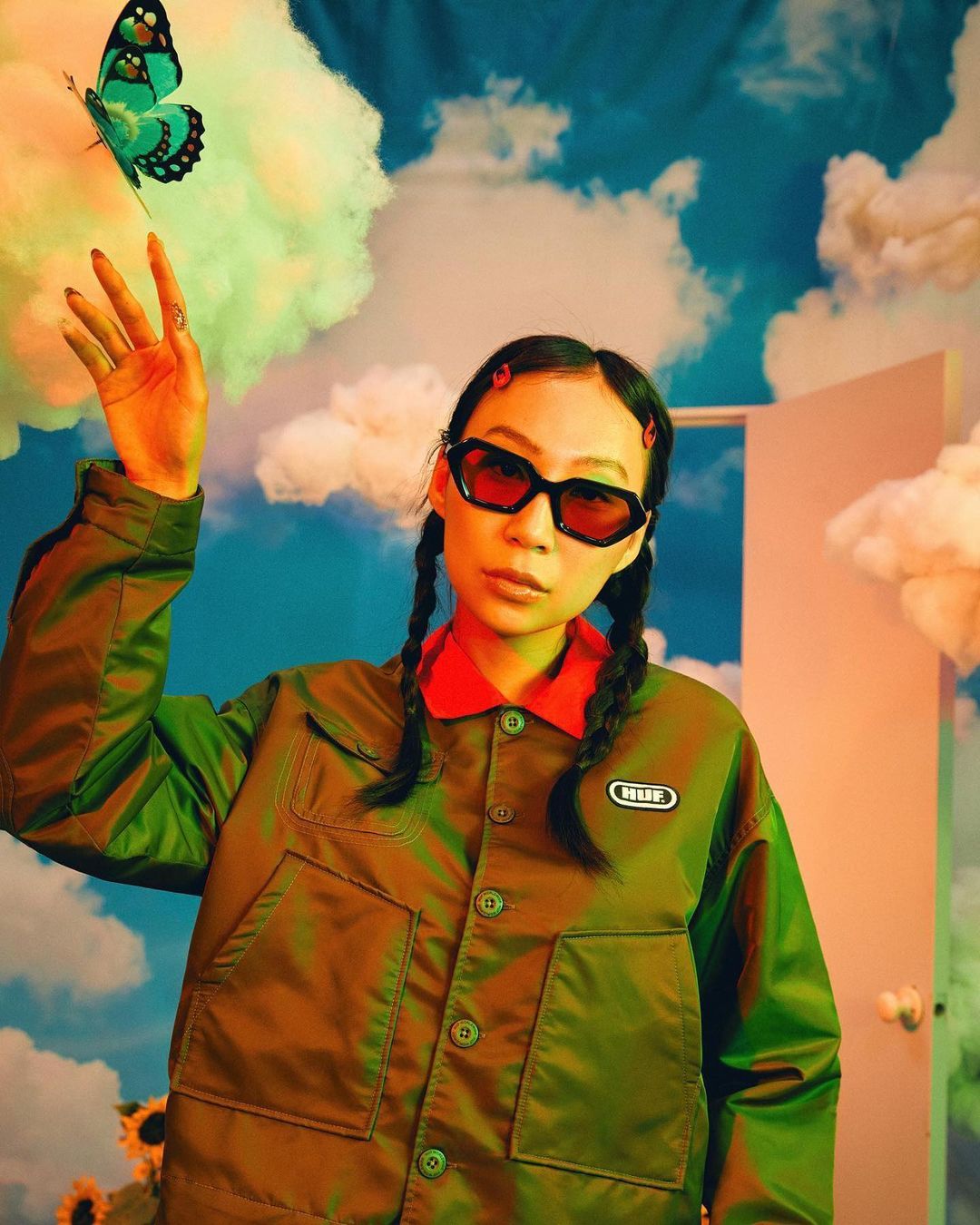 Portrait of a woman with braids, wearing red sunglasses and an olive green jacket, posing with a butterfly against a cloudy backdrop.