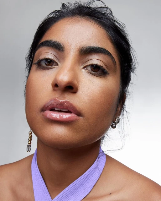 Close-up portrait of an adult woman with brown eyes and glossy lips wearing purple top and beaded earrings against a grey background.