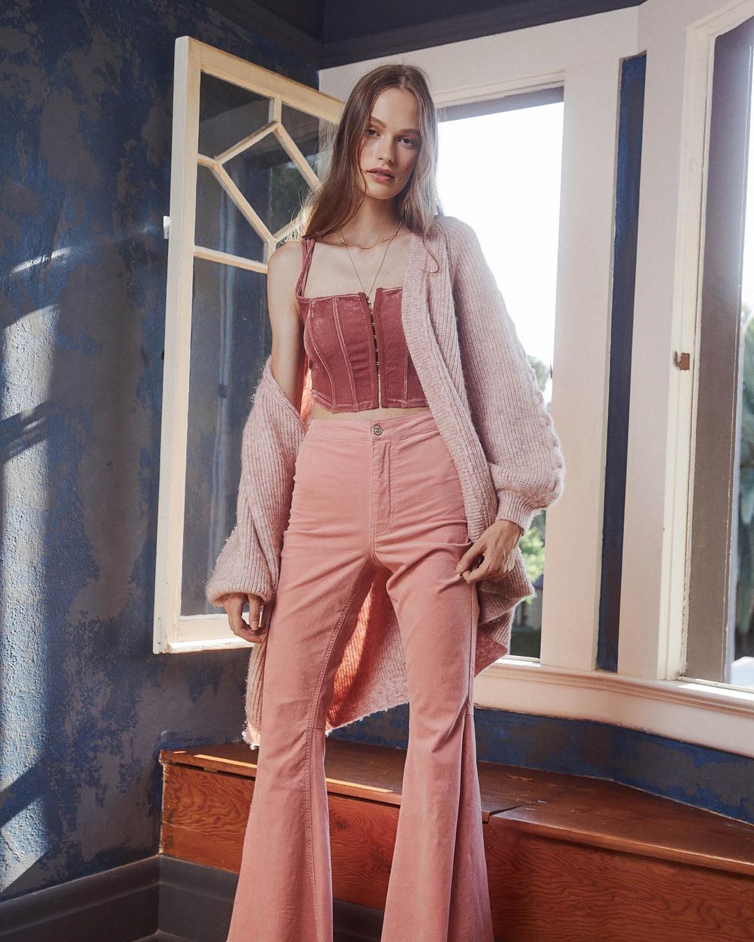 A woman in a pink corset top and flared pants stands indoors near a window, complemented by a cozy cardigan and soft, natural lighting.