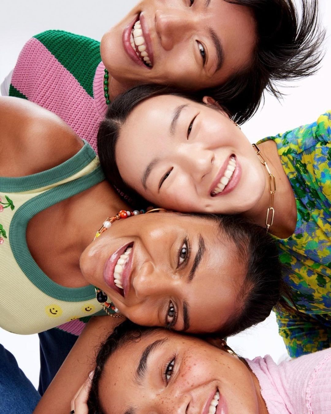 Four smiling multi-ethnic young adult faces close together in an eye-level, brightly lit studio shot, vibrant clothing, cheerful expressions.