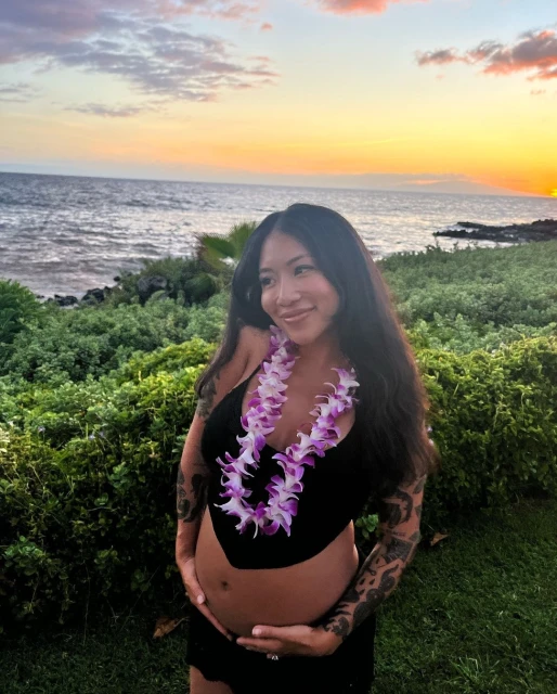 Pregnant woman poses outdoors at sunset in a black outfit and floral lei. Ocean and horizon visible in background. Tattoos on arms.