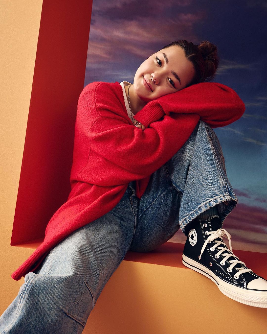 Woman in red cardigan and blue jeans sits in front of red and orange partitions with a cloudy sky backdrop wearing Converse sneakers.