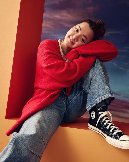 Woman in red cardigan and blue jeans sits in front of red and orange partitions with a cloudy sky backdrop wearing Converse sneakers.