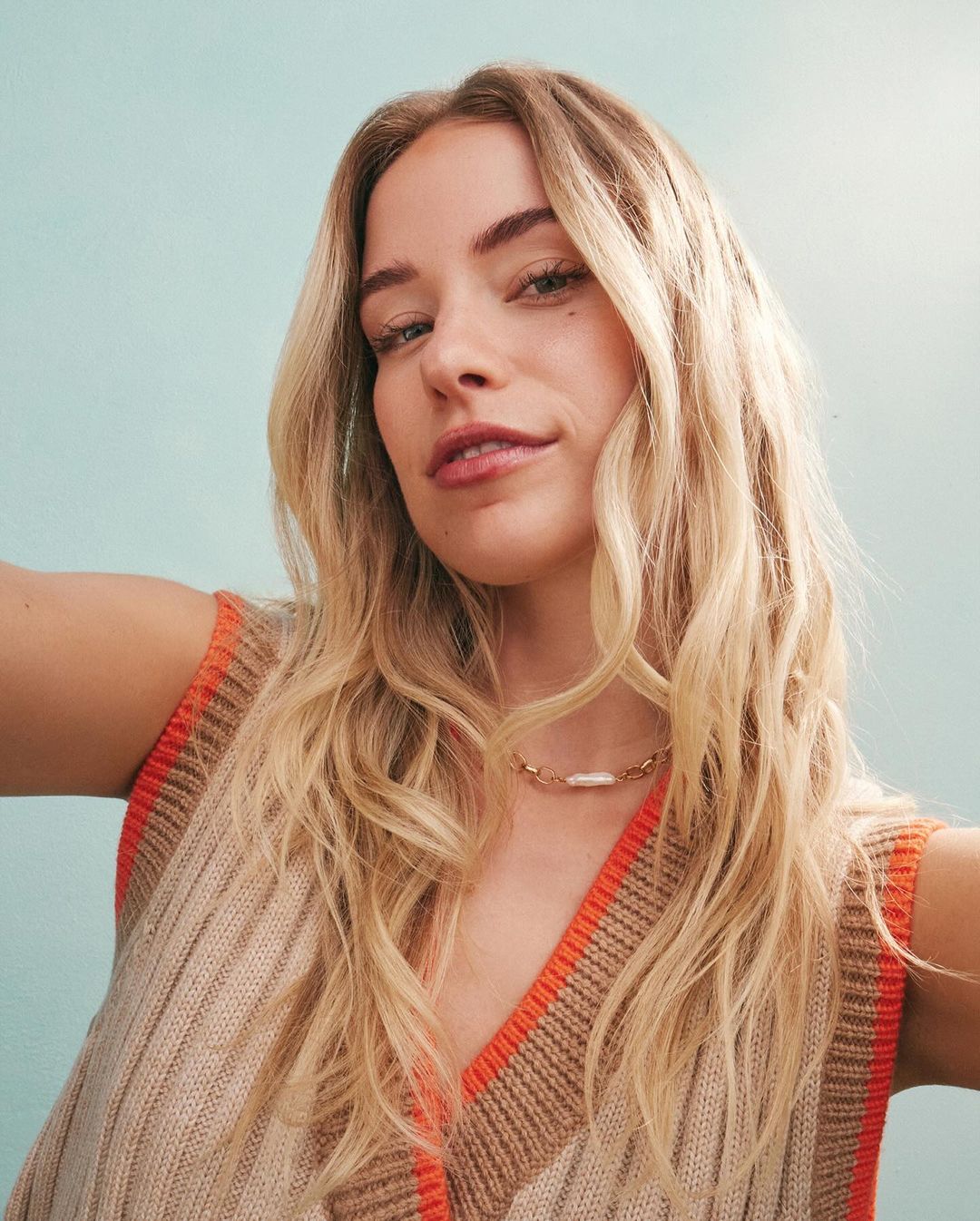 Close-up portrait of a blonde woman wearing a knitted vest and a gold necklace with a pearl accent, set against a light blue background.