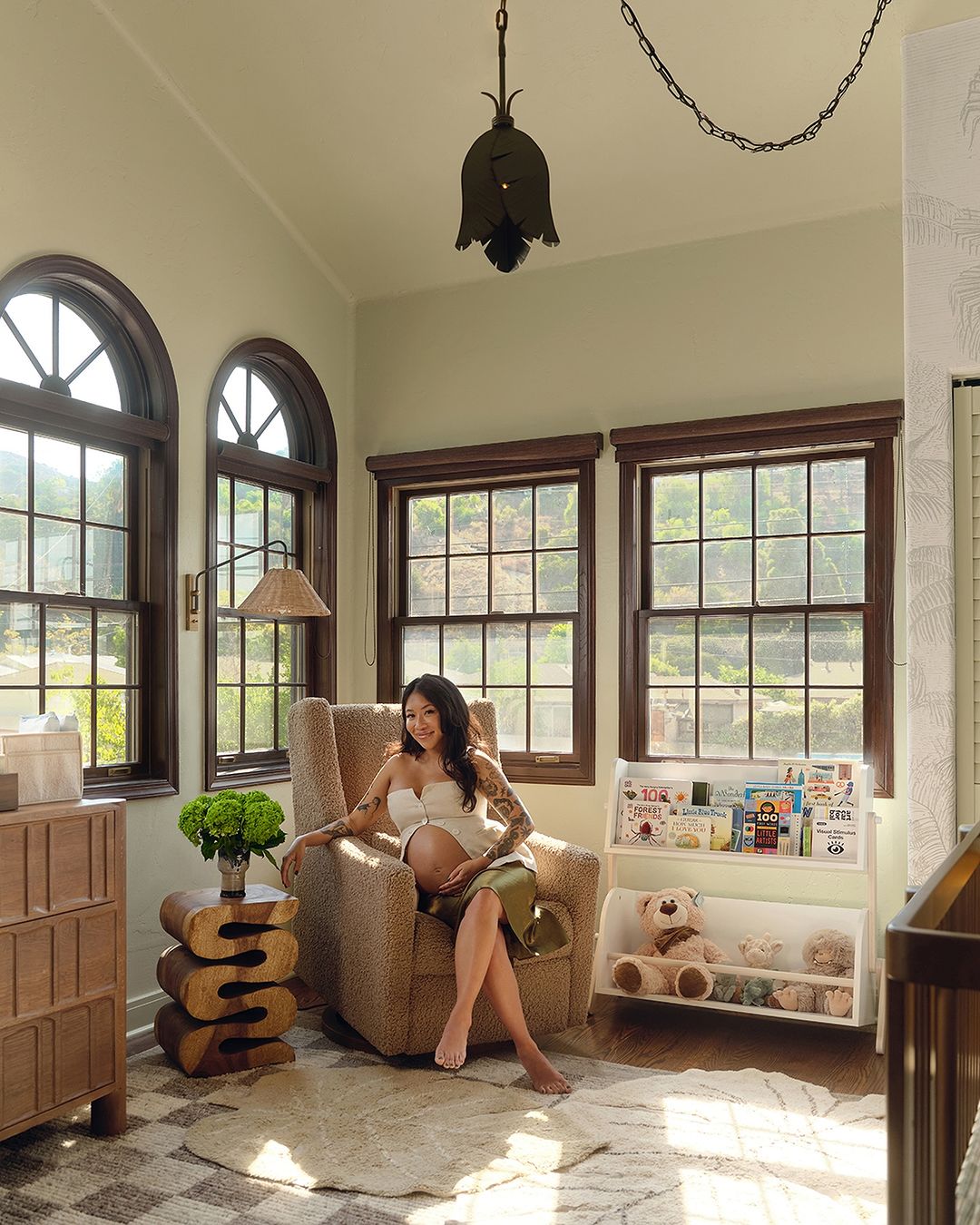 Pregnant woman with tattoos sits in a nursery with earth-tone colors and natural light from windows behind her, looking toward the camera.