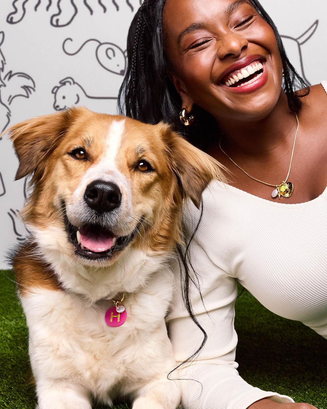 Portrait of a smiling woman posing with a fluffy dog wearing a pink tag with the letter H, against a backdrop with animal doodles.