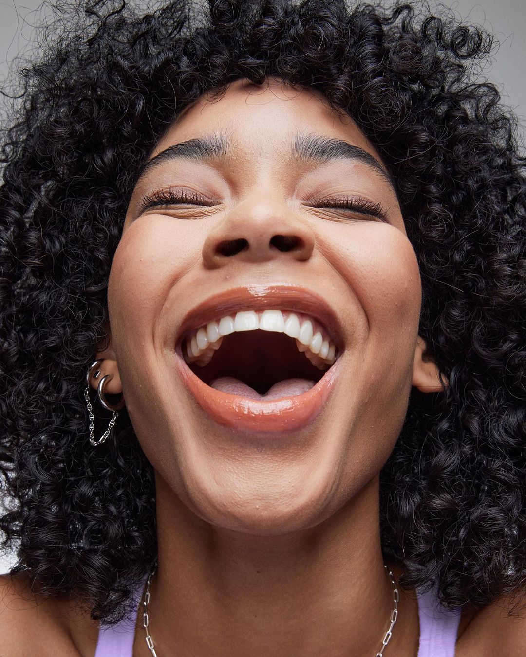 Close-up portrait of a smiling adult female with curly hair, showcasing a joyful expression and glossy lips in a bright, soft light.