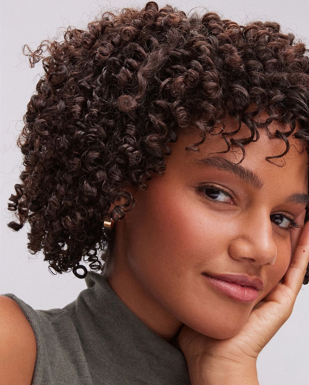 Close-up portrait of an adult woman with curly dark hair, wearing a sleeveless top and gold hoop earrings, resting her face on her hand.