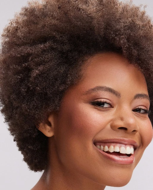 Close-up portrait of a smiling adult woman with brown skin and voluminous afro hairstyle against a neutral grey background.