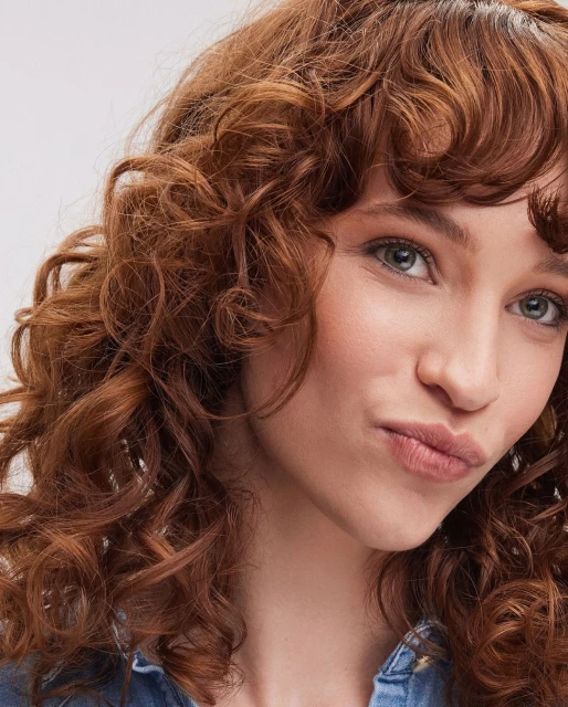Close-up portrait of an adult woman with curly red hair, wearing a blue denim top against a neutral background, focusing on her face and hair.