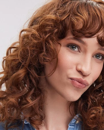 Close-up portrait of an adult woman with curly red hair, wearing a blue denim top against a neutral background, focusing on her face and hair.