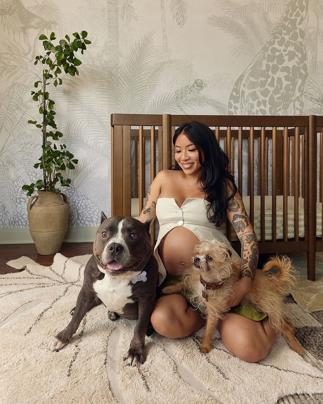 Pregnant woman poses indoors with two dogs; she is kneeling on an area rug in front of a dark wood baby crib. A houseplant is visible.