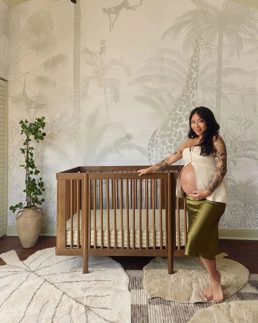 Pregnant woman in a neutral-toned top and green skirt stands in a nursery with a wooden crib, tropical-themed wallpaper, and leaf-shaped rugs.