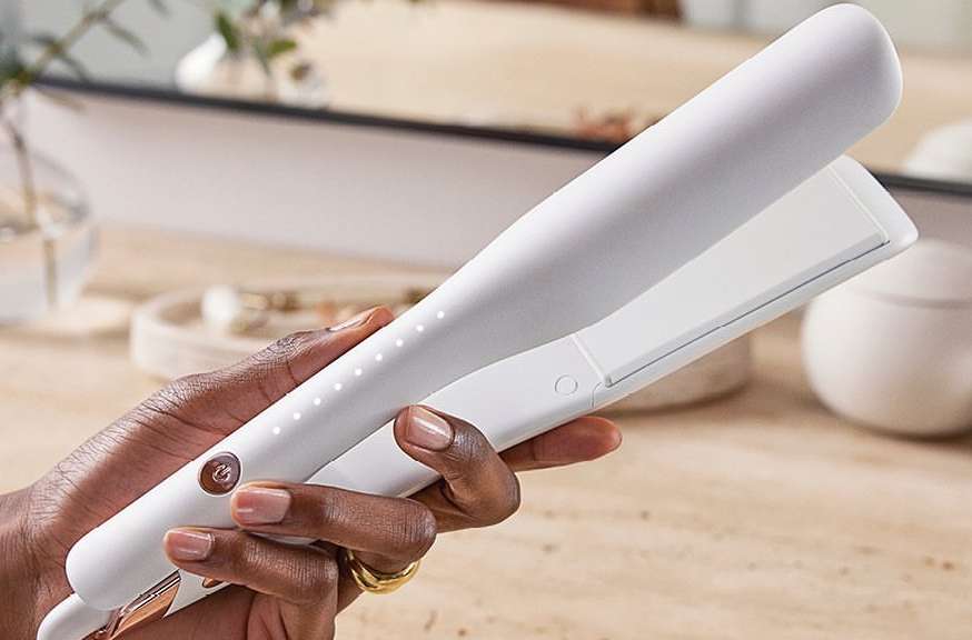 Adult hand holds a white and gold hair straightener in an indoor setting with a mirror in the background. Beauty product close-up.