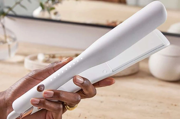 Adult hand holds a white and gold hair straightener in an indoor setting with a mirror in the background. Beauty product close-up.