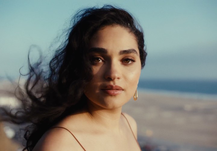 Close-up of a woman with dark curly hair and gold earrings, posing against a background of beach and blue sky in outdoor setting.