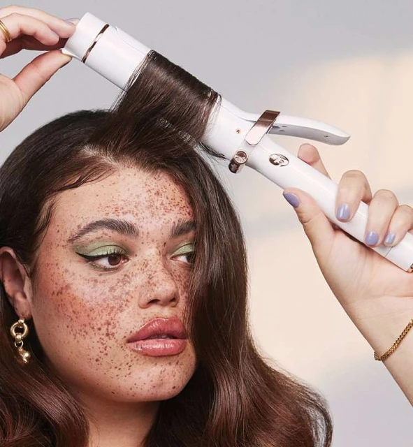 Adult female curling hair with a white and rose-gold curling iron in a beauty shoot; neutral background; light lilac nail polish visible.