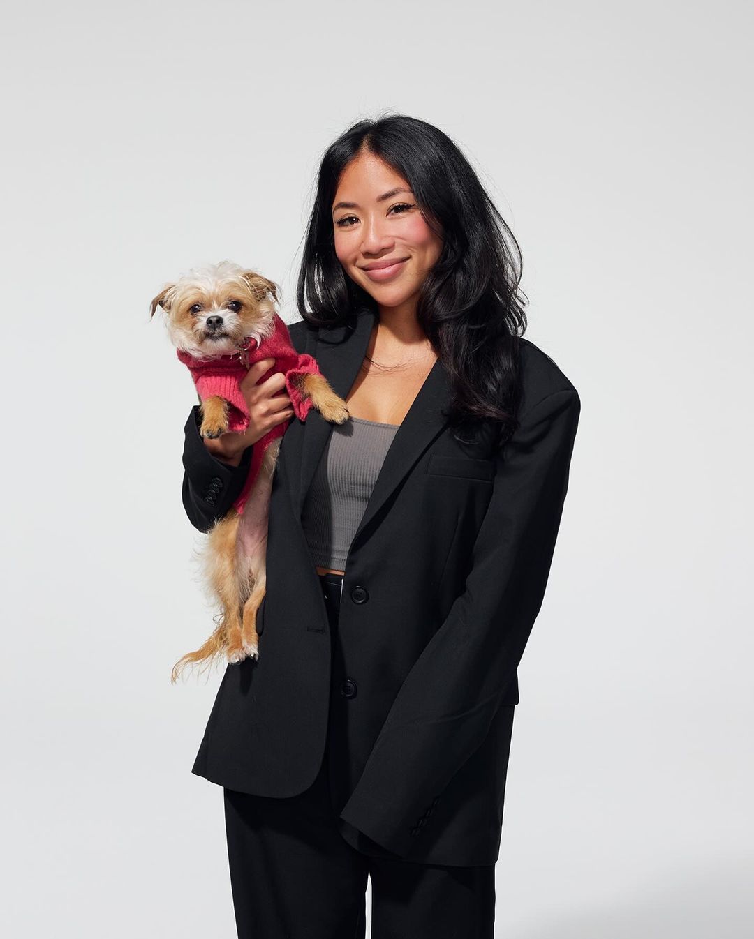 Asian woman in a black blazer holding a small dog wearing a pink sweater in a photo studio with a white background. Fashion editorial.