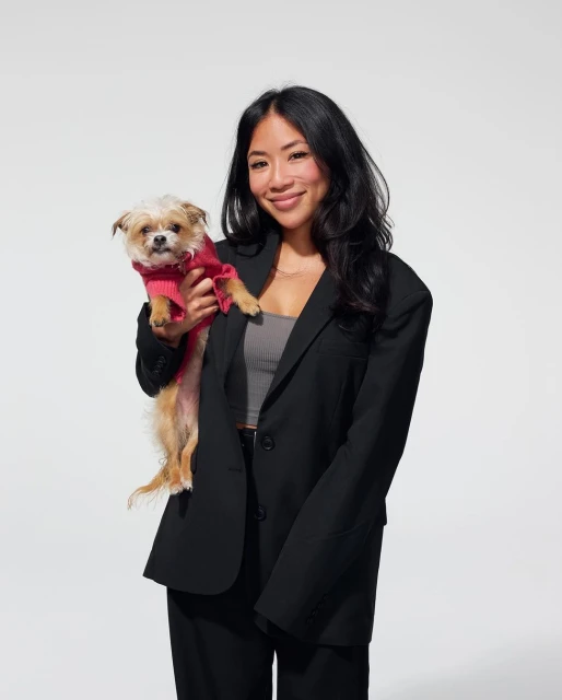 Asian woman in a black blazer holding a small dog wearing a pink sweater in a photo studio with a white background. Fashion editorial.