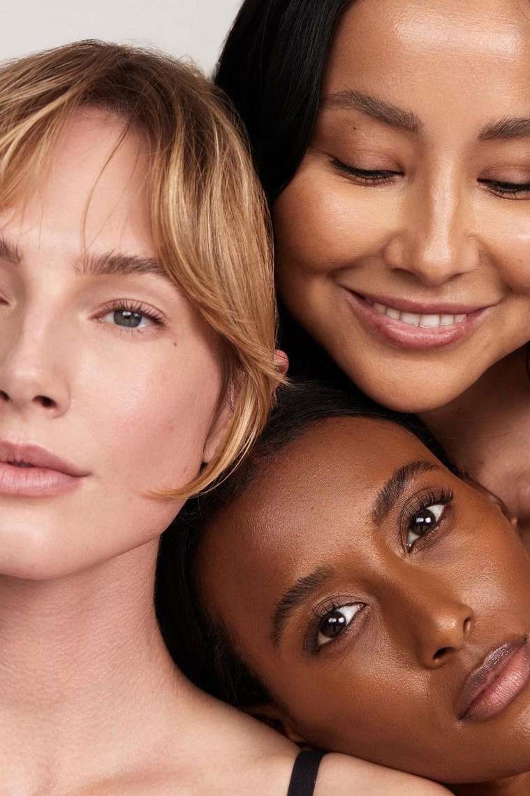 Beauty portrait of three diverse young women with different skin tones and hairstyles posing closely together in a brightly lit studio.