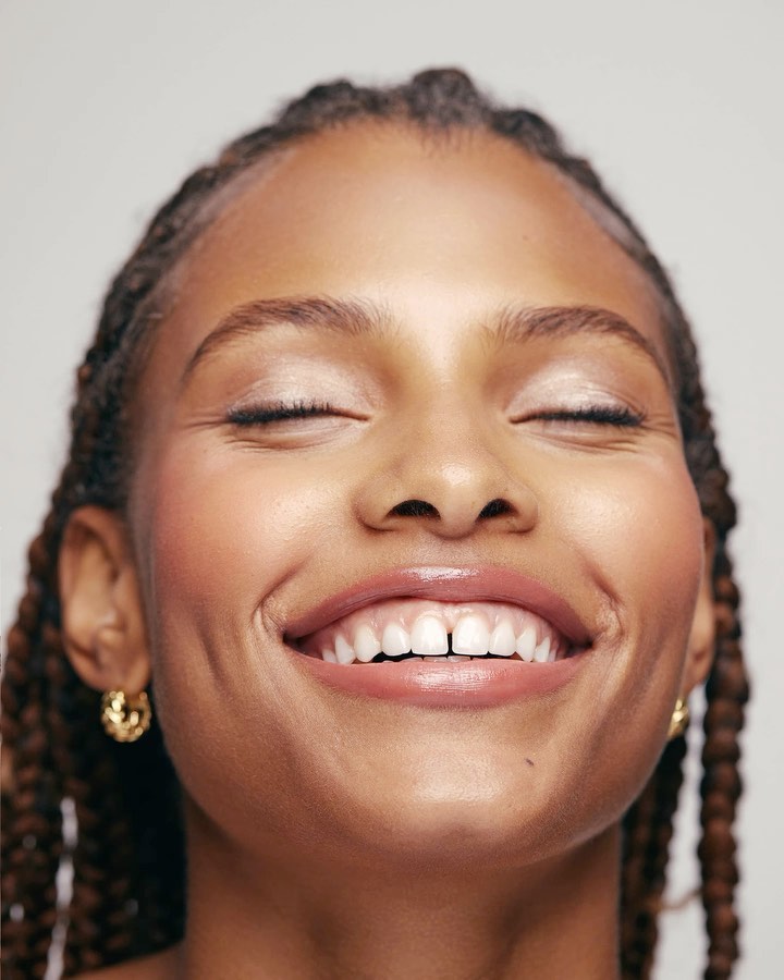 Close-up portrait of a smiling Black woman with braided hair, dewy makeup, and gold earrings against a bright neutral backdrop.