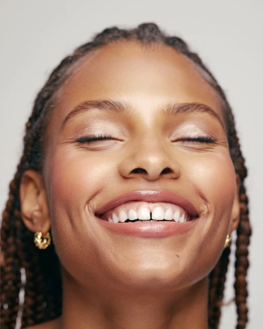 Close-up portrait of a smiling Black woman with braided hair, dewy makeup, and gold earrings against a bright neutral backdrop.