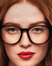 Close-up portrait of a woman with red hair, freckles, and red lipstick wearing black eyeglasses.