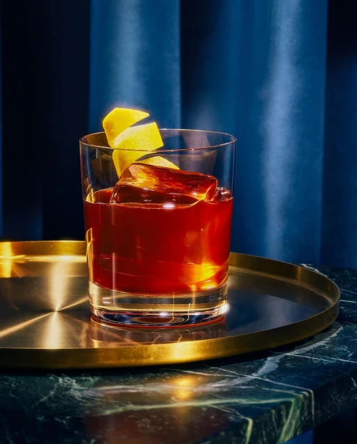 A glass filled with a reddish cocktail and ice, garnished with a lemon twist, sits on a gold tray against a dark blue backdrop.