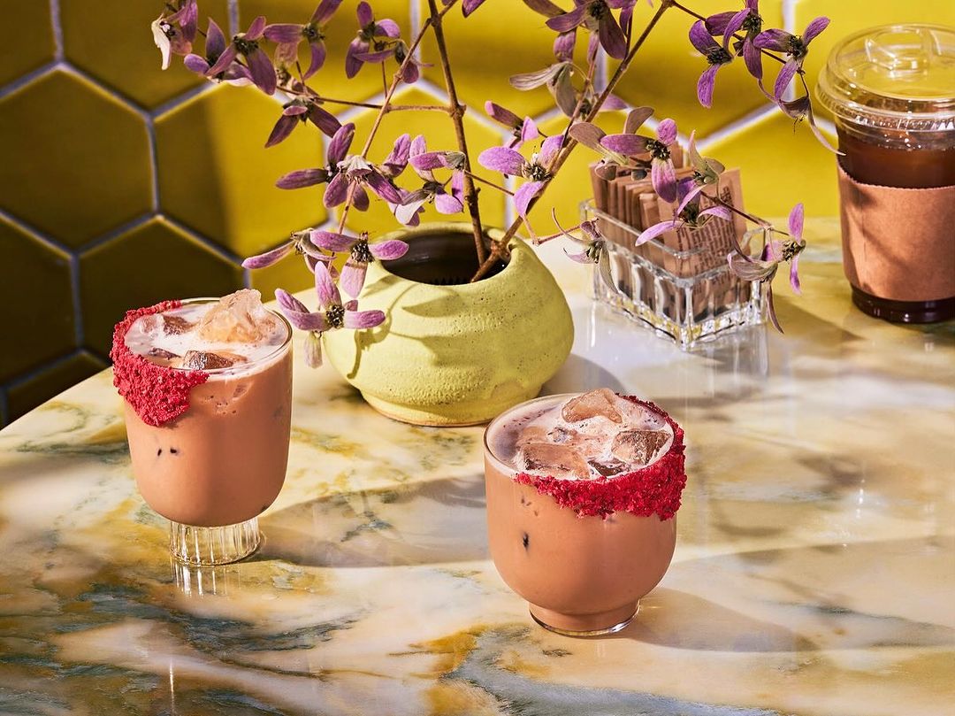 Two iced mocha cocktails garnished with red sprinkles, alongside a flower arrangement, and a plastic cup of coffee on a marble table.