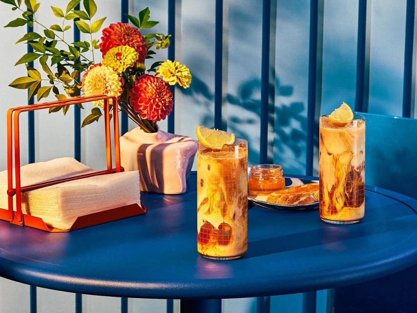 Two iced coffees with lemon slices sit on a blue table next to flowers, napkins, and jam. Soft lighting enhances the cool, refreshing drink.