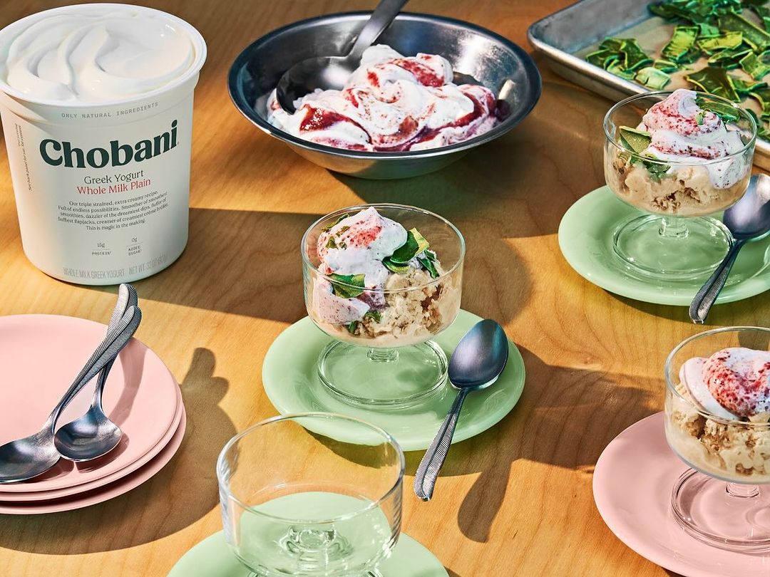 Chobani Greek yogurt with dessert bowls, plates, and spoons on a wooden table, showcasing a food photography still life.