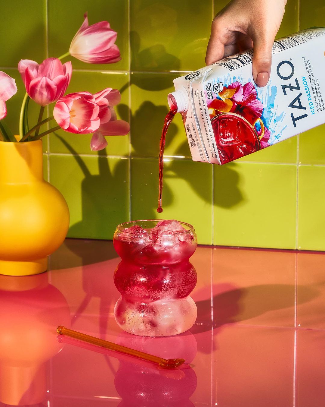 Iced tea is poured from a carton into a clear glass with ice, alongside pink tulips in a yellow vase and a pink stirring stick.