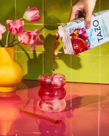 Iced tea is poured from a carton into a clear glass with ice, alongside pink tulips in a yellow vase and a pink stirring stick.