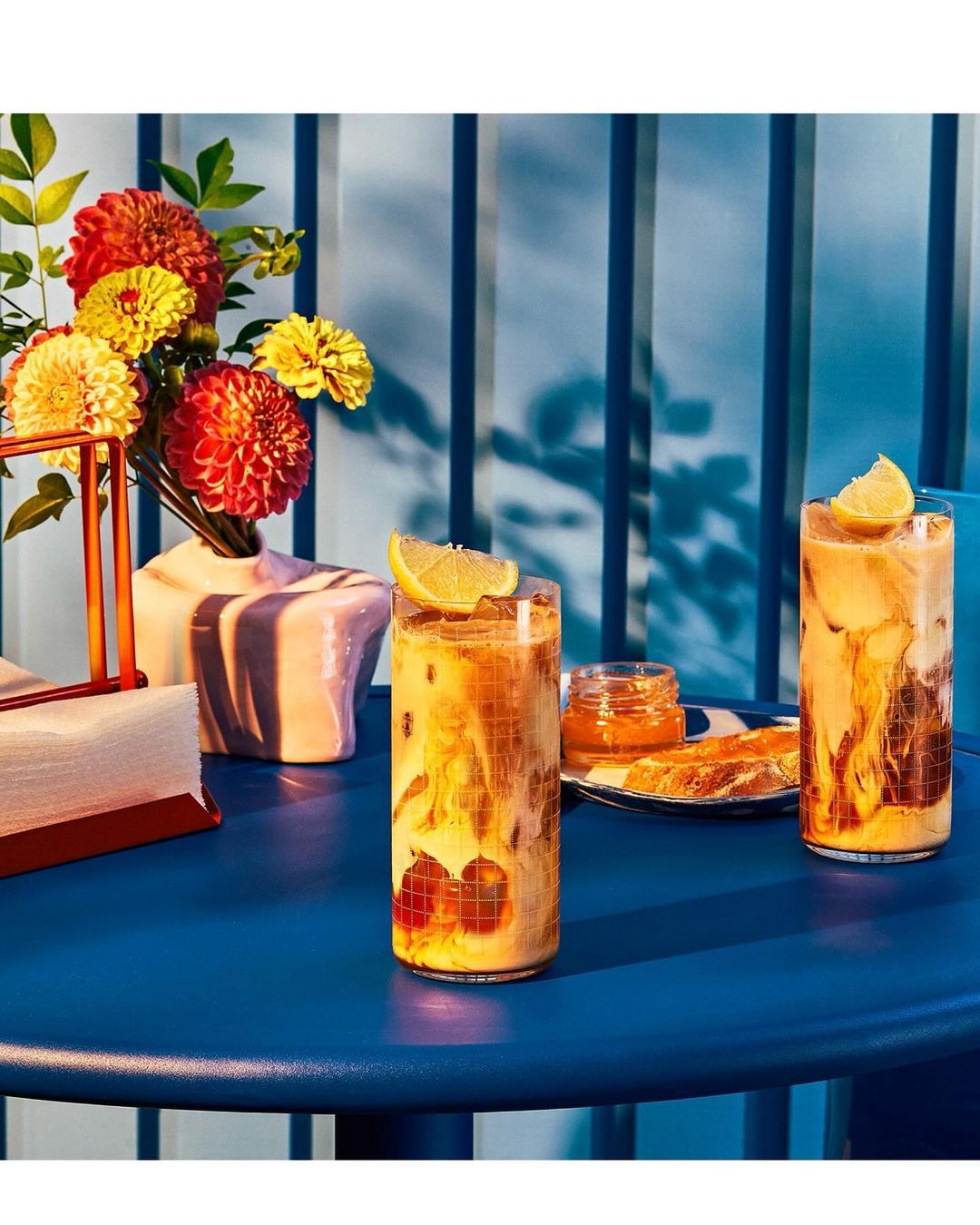 Two iced beverages on a blue table, accented by a bouquet of colorful flowers, a jar of jam, and blue striped background.