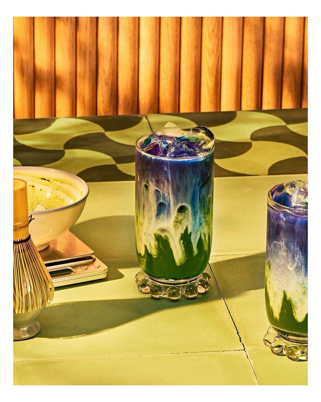 Two colorful, layered matcha lattes in tall glasses, accented with a wavy green background and a decorative wooden screen.