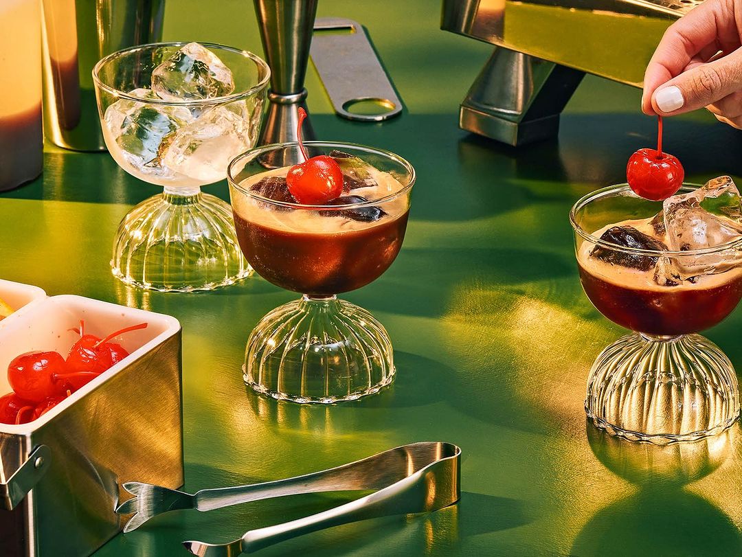 Three cocktails with cherries and ice, arranged on a green surface, with a cocktail mixing kit, tongs, and a metal dish.