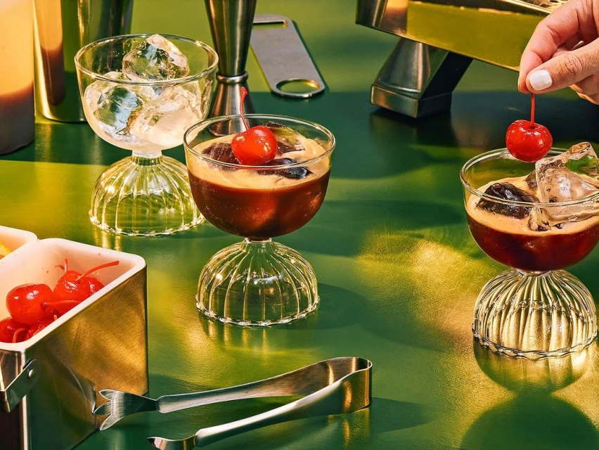 Three cocktails with cherries and ice, arranged on a green surface, with a cocktail mixing kit, tongs, and a metal dish.