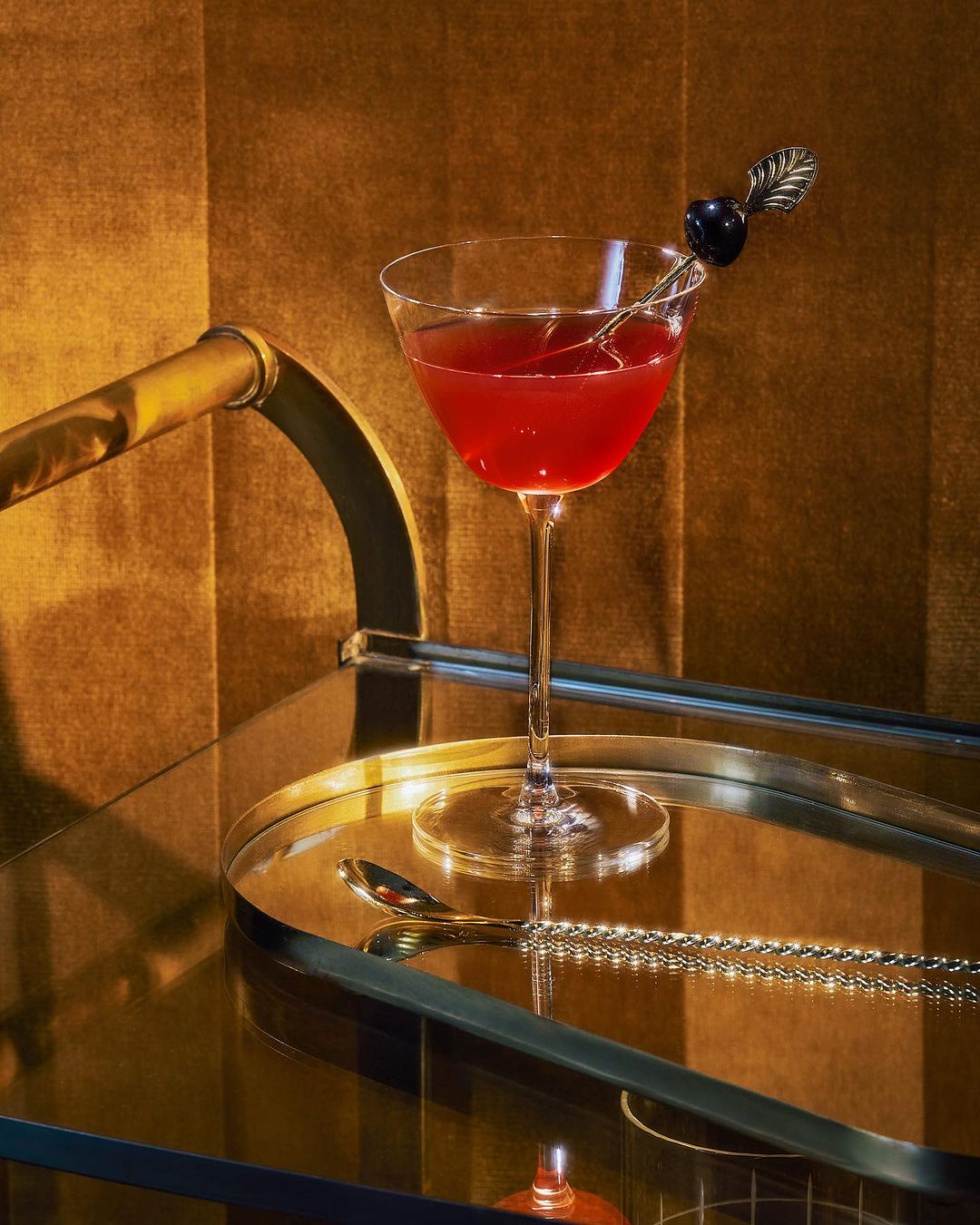 Elegant cocktail glass with dark red liquid, garnished with a black cherry, sits on a mirrored bar cart against a golden backdrop.