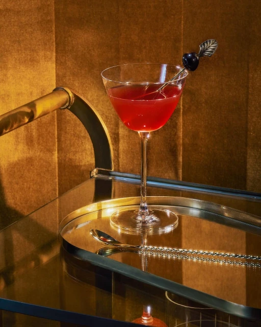 Elegant cocktail glass with dark red liquid, garnished with a black cherry, sits on a mirrored bar cart against a golden backdrop.