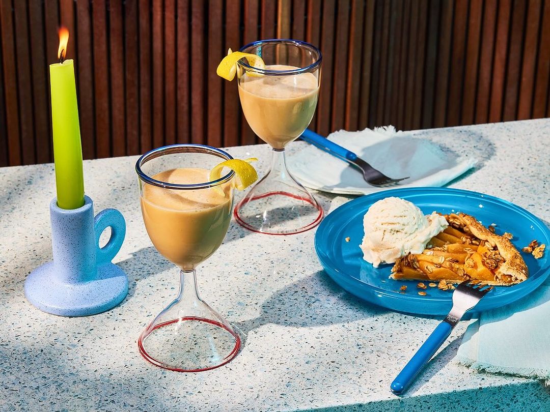 Still life of two dessert cocktails with lemon twists, slice of pie with ice cream, and a green candle on a white textured surface.