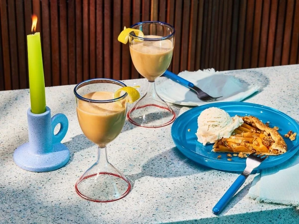 Still life of two dessert cocktails with lemon twists, slice of pie with ice cream, and a green candle on a white textured surface.