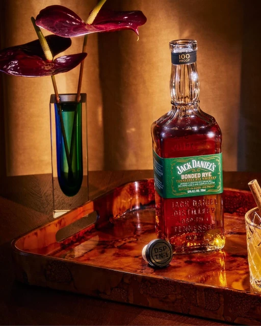 Bottle of Jack Daniel's Bonded Rye whiskey on a brown tray with a vase of red flowers and a cocktail in a glass.