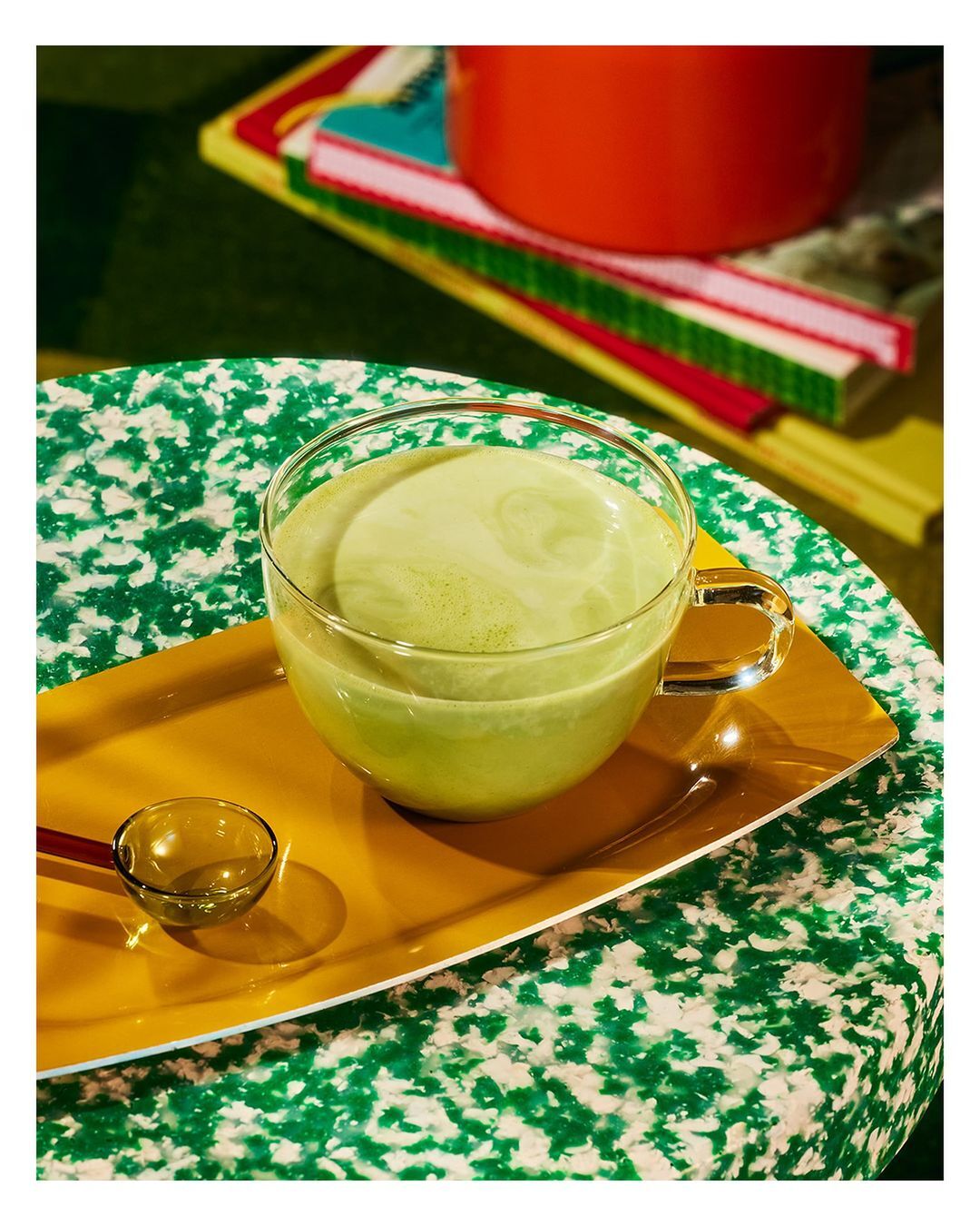 A clear glass cup filled with green matcha latte sits on a yellow tray, with books and an orange object blurred behind it.