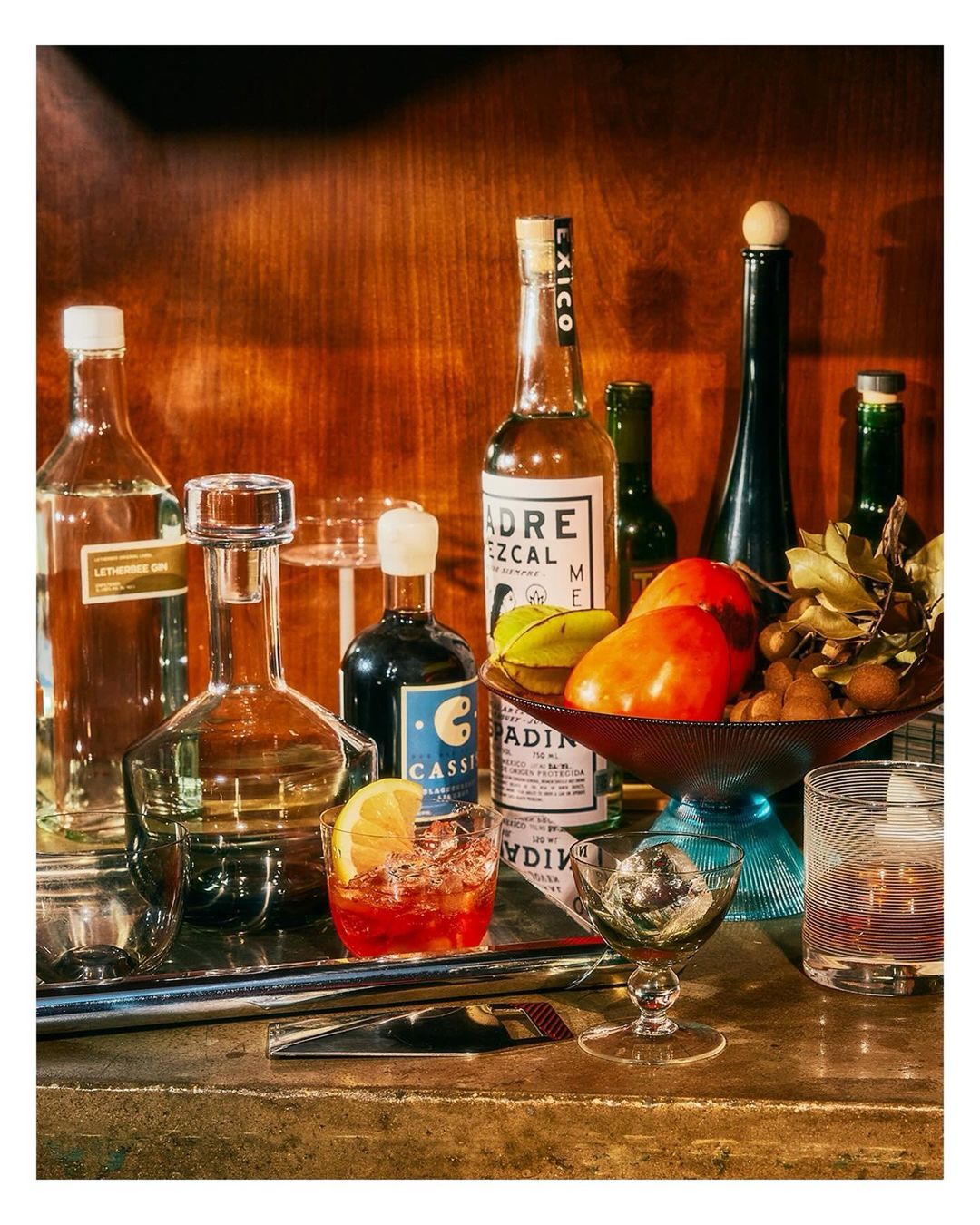 Still life photograph of cocktails, bottles, and fruit arranged on a bar surface against a wooden backdrop in warm, inviting lighting.