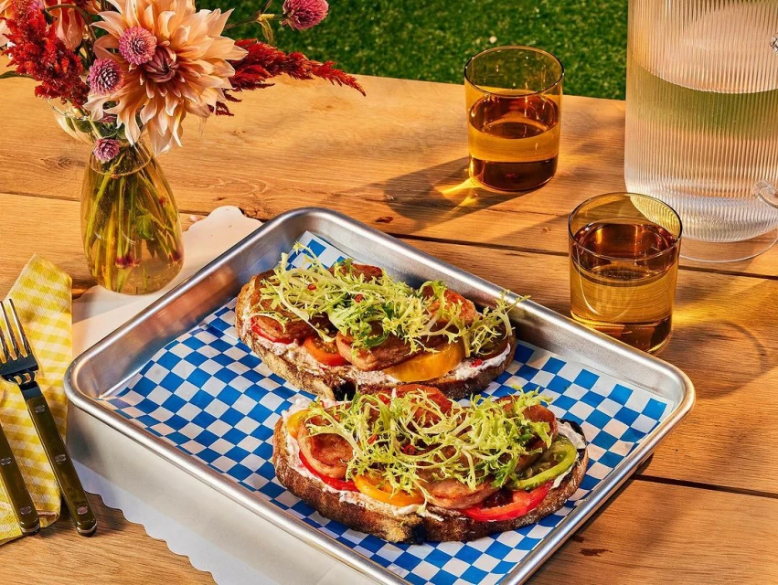 Two open-faced sandwiches with sliced sausage, heirloom tomatoes and greens on a tray with beverage and flowers, outdoor setting.